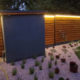 EuNike Architektura Krajobrazu Eunika Wiatr - Oświetlony ogród z drewnianym ogrodzeniem, ścieżką z kamieni i roślinnością ozdobną. Widoczne lampy LED nadające ciepłe światło. Elementy małej architektury.