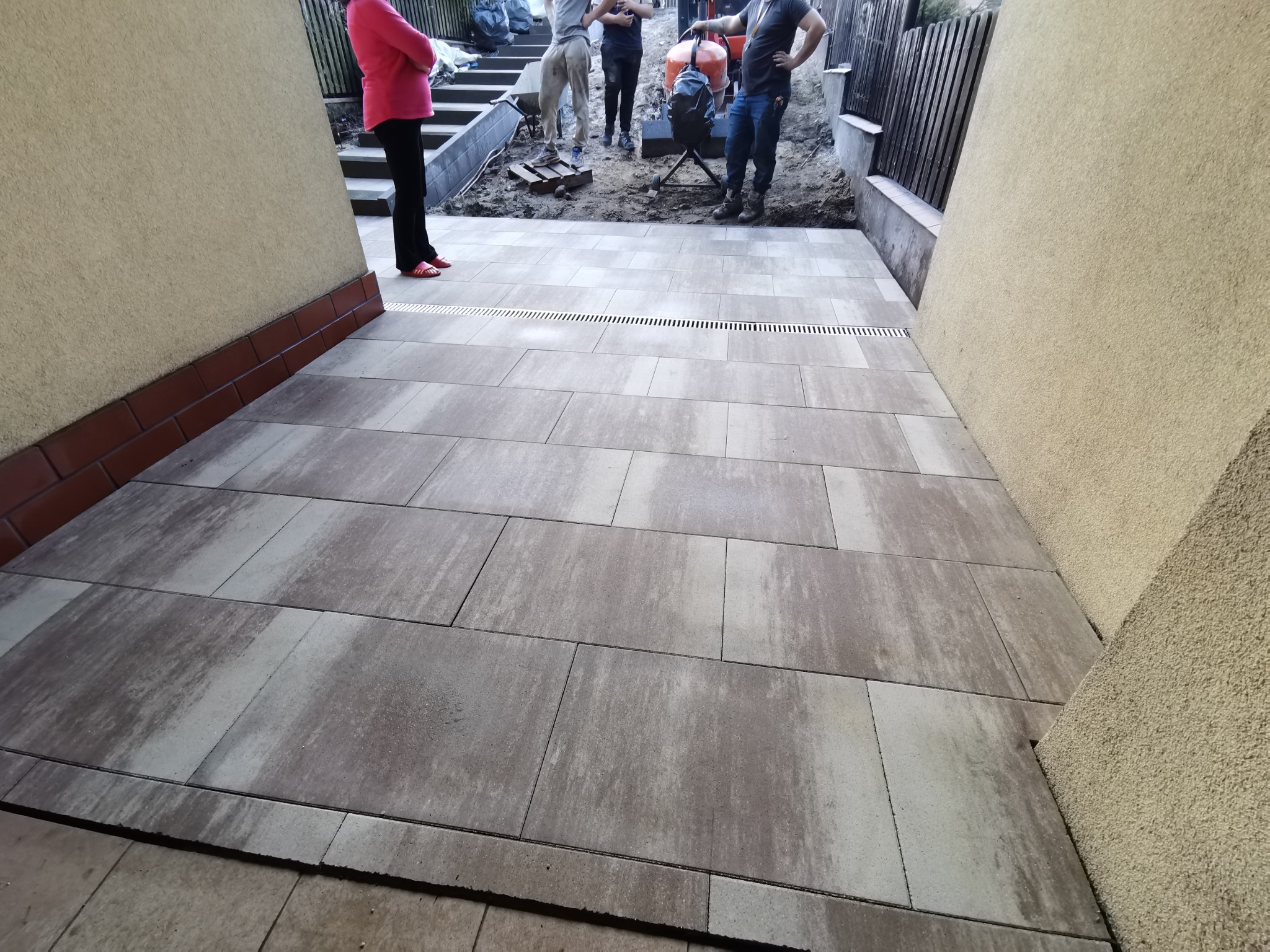 Ułożona, prostokątna kostka brukowa w odcieniach szarości z widocznym odpływem liniowym, ekipa montażowa w tle z betoniarką.