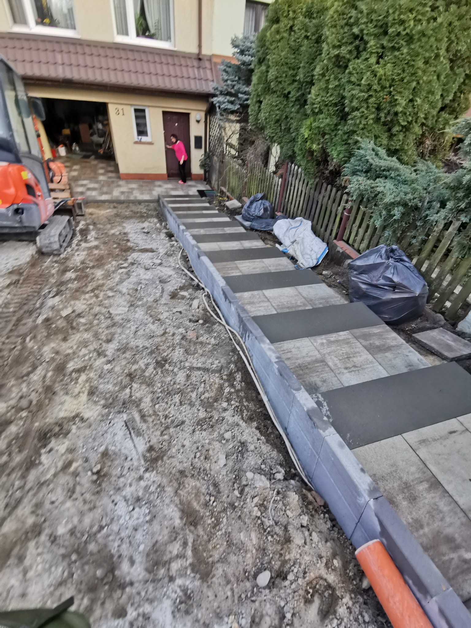 Układanie stopni z kostki brukowej przed domem jednorodzinnym, widoczny mini-koparka i kobieta w różowej bluzce przy drzwiach wejściowych.