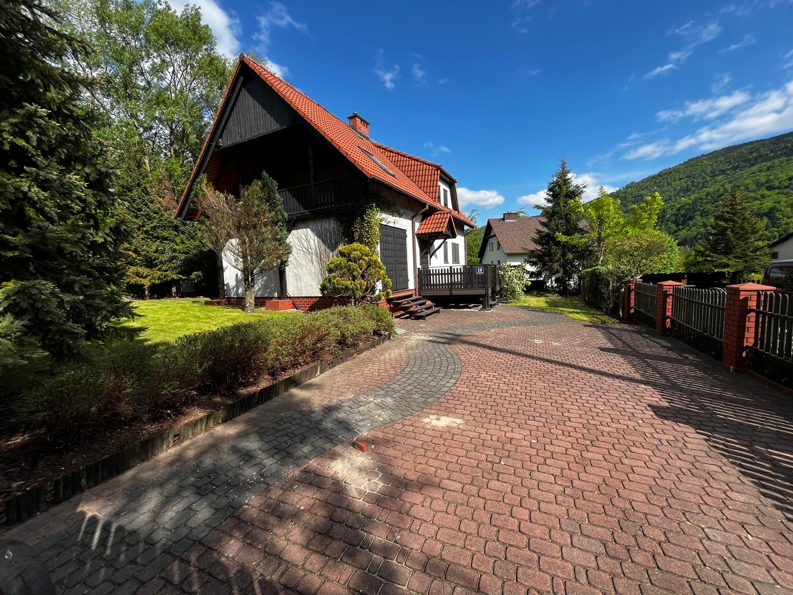 Czerwona kostka brukowa prowadzi do domu z ogrodem. Widok na posesję w słoneczny dzień, z elementami zieleni i górskim krajobrazem w tle.