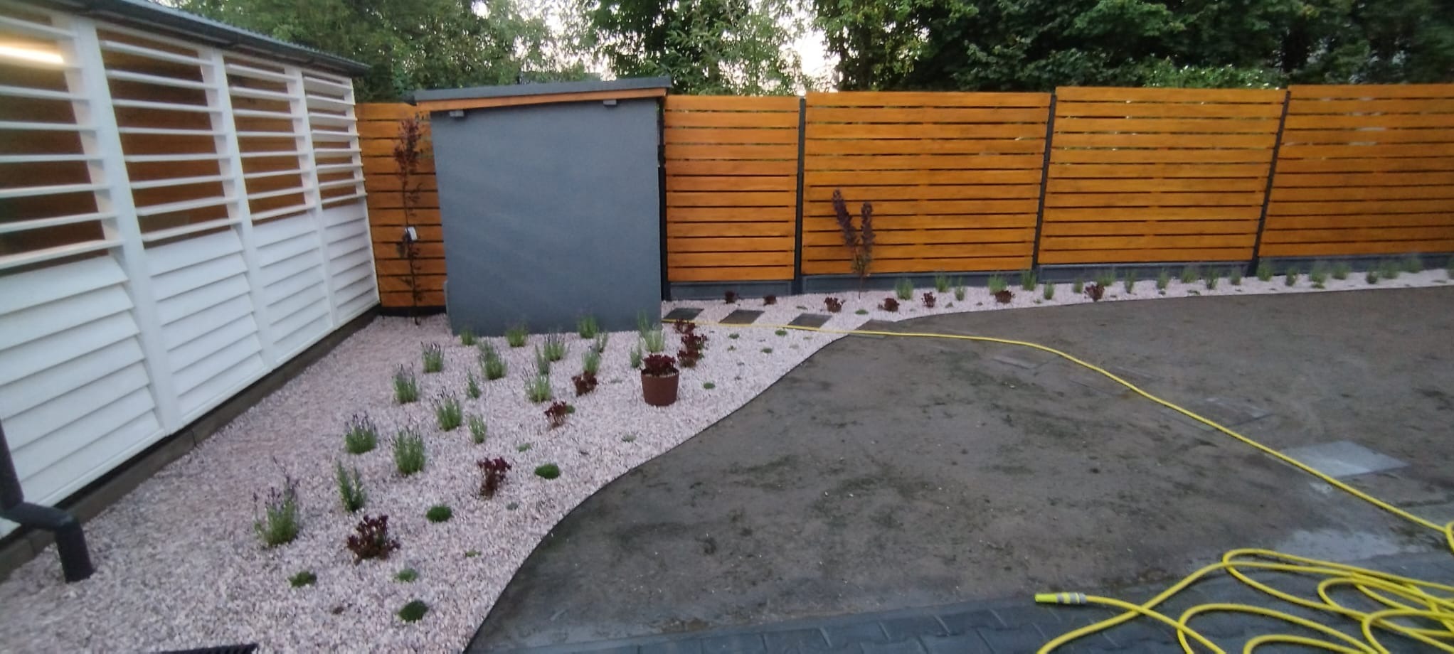 Nowoczesny ogród po realizacji: drewniane ogrodzenie, biała zabudowa, rabaty z kamyków i roślin, żółty wąż ogrodowy na ziemi. Minimalistyczny design.