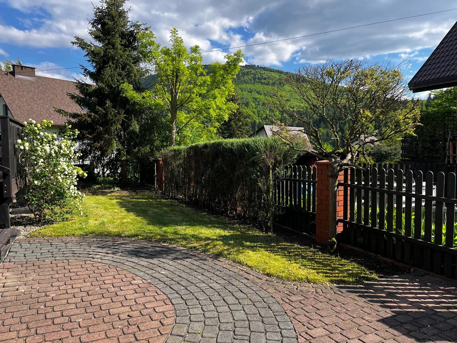 Ogród z alejką z kostki, żywopłotem i czarnym ogrodzeniem na tle zalesionego wzgórza pod błękitnym niebem z chmurami. Słoneczny dzień.