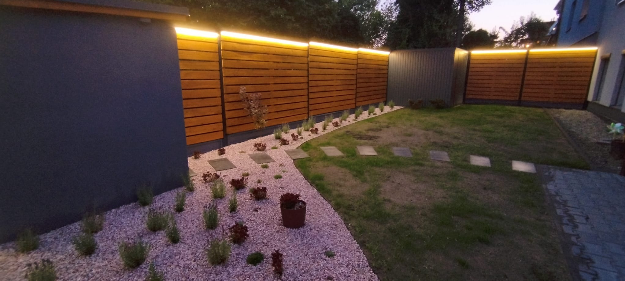 Nowoczesny ogród po zmroku: podświetlony drewniany płot, ścieżka z kamyczków i płyt, trawnik. Minimalistyczny design z subtelnym oświetleniem.