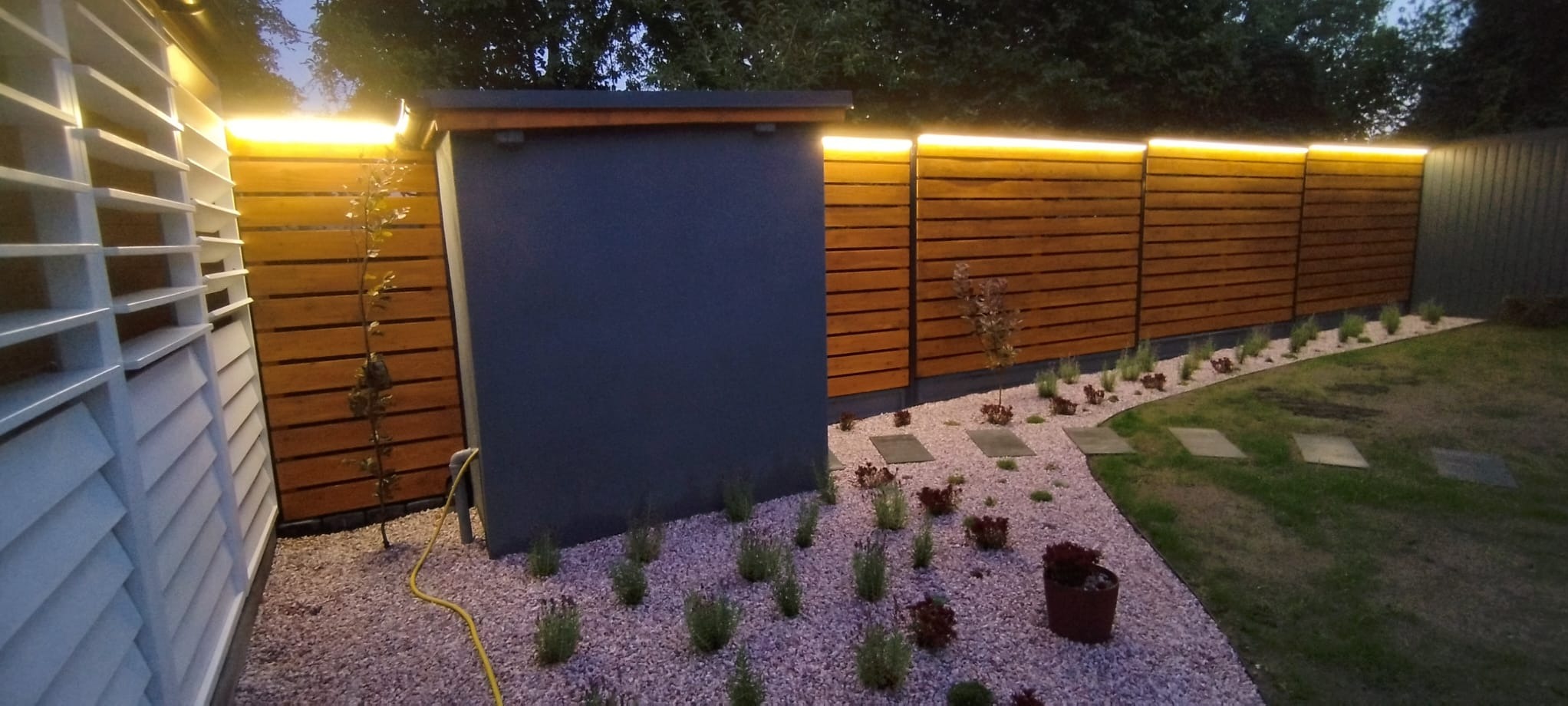 Oświetlony ogród z drewnianym ogrodzeniem, ścieżką z kamieni i roślinnością ozdobną. Widoczne lampy LED nadające ciepłe światło. Elementy małej architektury.