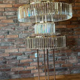 Trójpoziomowy, kryształowy żyrandol na złotych nóżkach, ustawiony na drewnianej podłodze na tle ściany z czerwonej cegły.