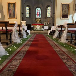 Ale ładnie - Uroczysta dekoracja kościoła na ślub: czerwony dywan, sztuczna trawa z białymi kwiatami wzdłuż ławek, białe tiulowe kokardy z zielonymi gałązkami na ławkach, ołtarz w tle.