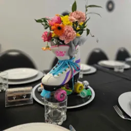 Dekoracja stołu z rolką w stylu retro jako wazonem na kwiaty, otoczona kolorowymi kółkami i kasetą magnetofonową 'Biblia dla dzieci', na czarnym obrusie.