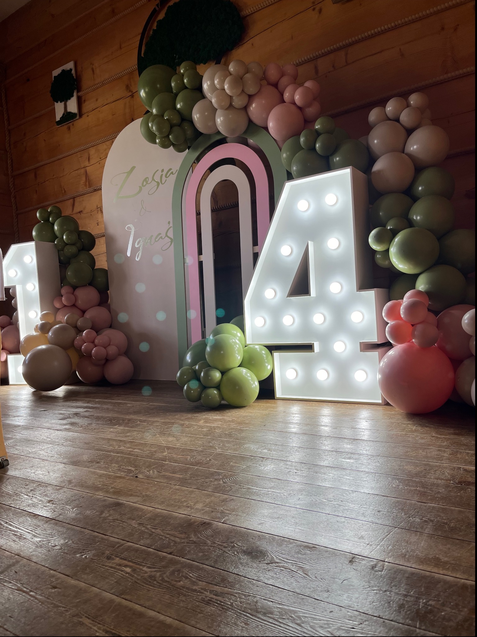Dekoracja urodzinowa z podświetlanymi cyframi '1' i '4', otoczona girlandami z pastelowych balonów w odcieniach zieleni, beżu i różu, na tle drewnianej ściany i podłogi.