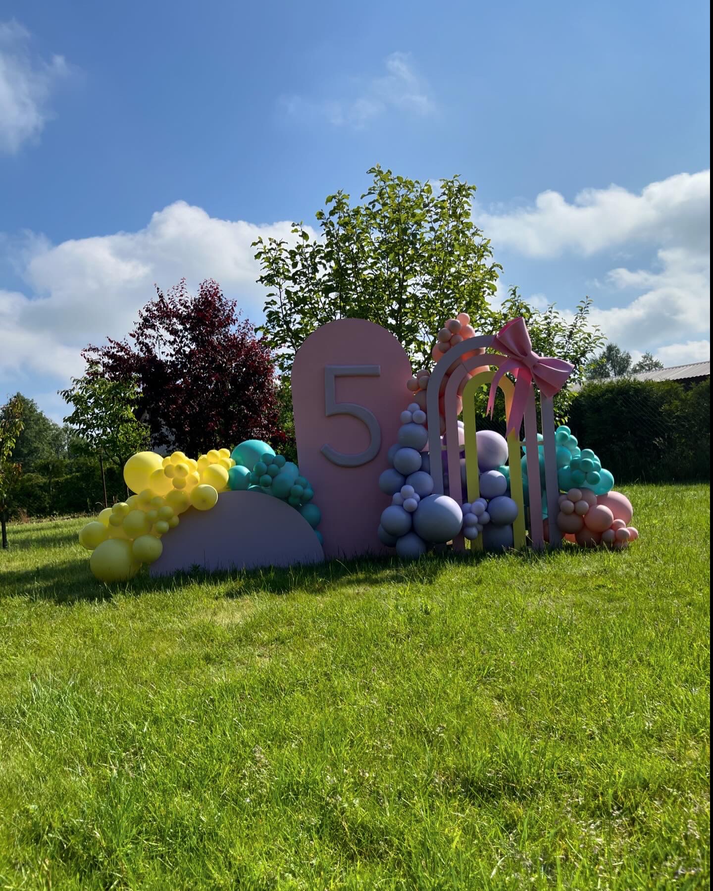 Dekoracja urodzinowa na trawie z pastelowymi balonami, łukiem i cyfrą '5' w słoneczny dzień.