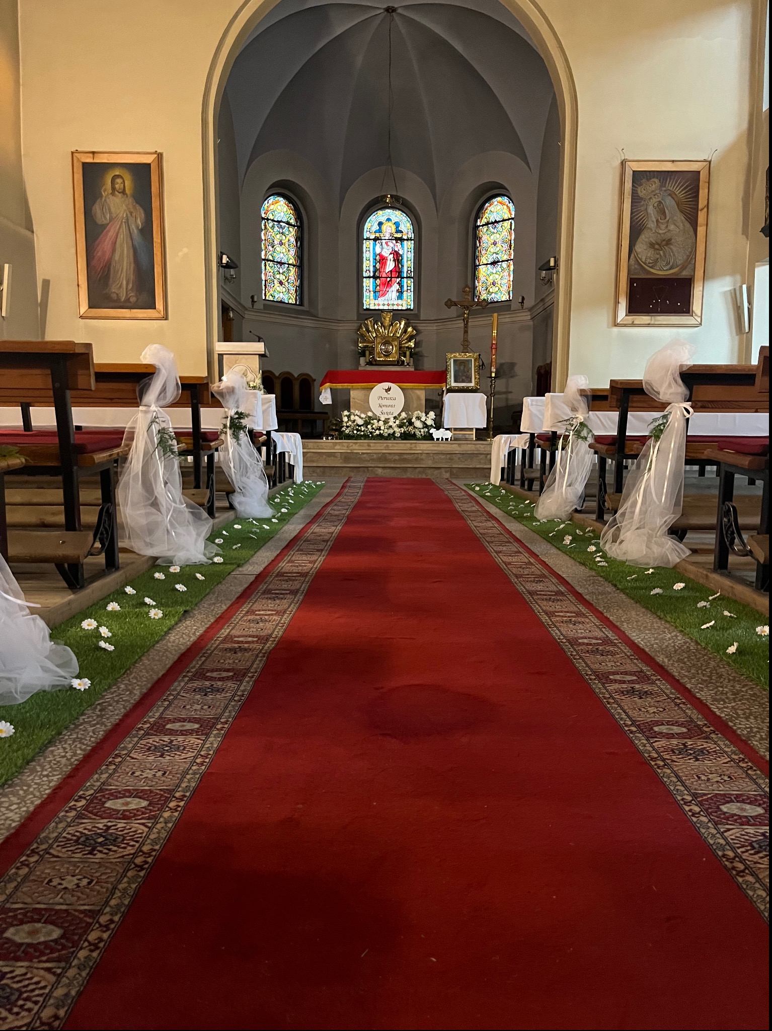 Uroczysta dekoracja kościoła na ślub: czerwony dywan, sztuczna trawa z białymi kwiatami wzdłuż ławek, białe tiulowe kokardy z zielonymi gałązkami na ławkach, ołtarz w tle.