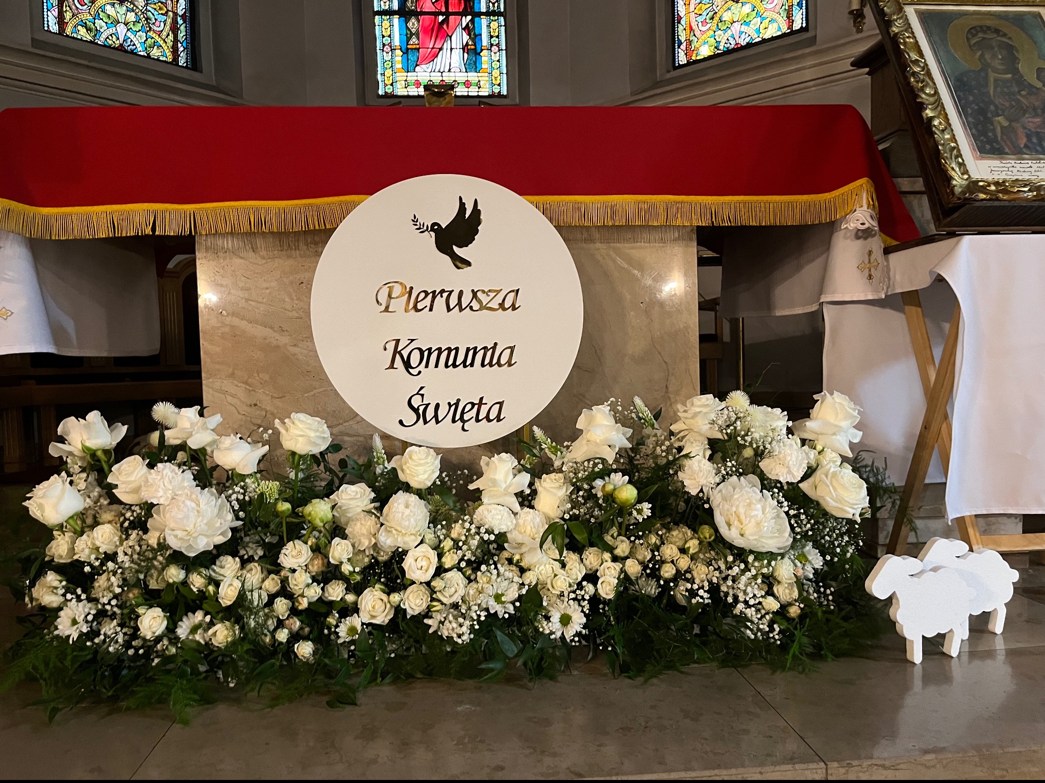 Dekoracja ołtarza na Pierwszą Komunię Świętą: białe kwiaty, okrągły napis z gołębiem, drewniane owieczki, kościół w tle.