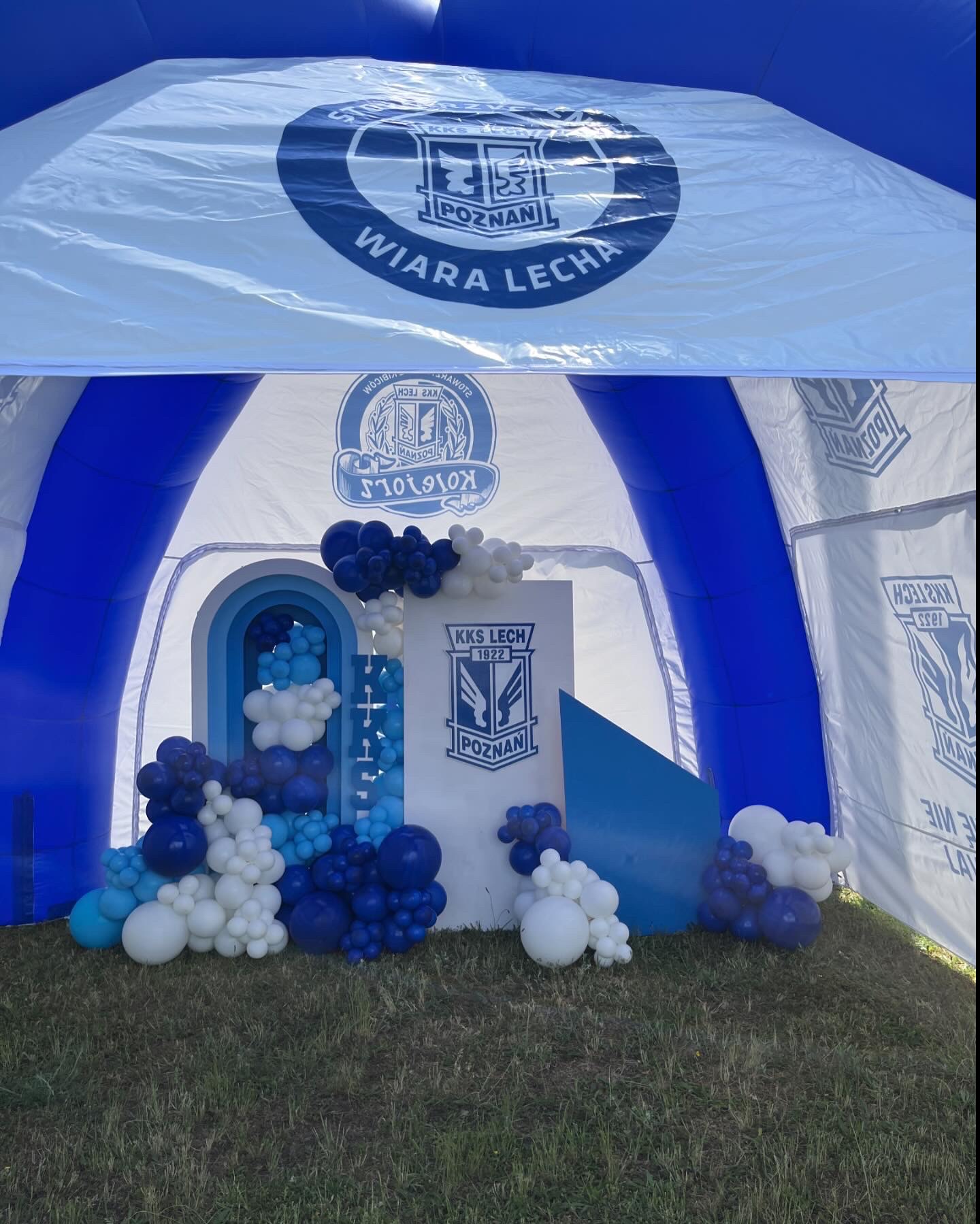 Dekoracja balonowa w odcieniach bieli i niebieskiego, z logo klubu sportowego KKS Lech Poznań, umieszczona wewnątrz namiotu z brandingiem klubowym, na trawie.