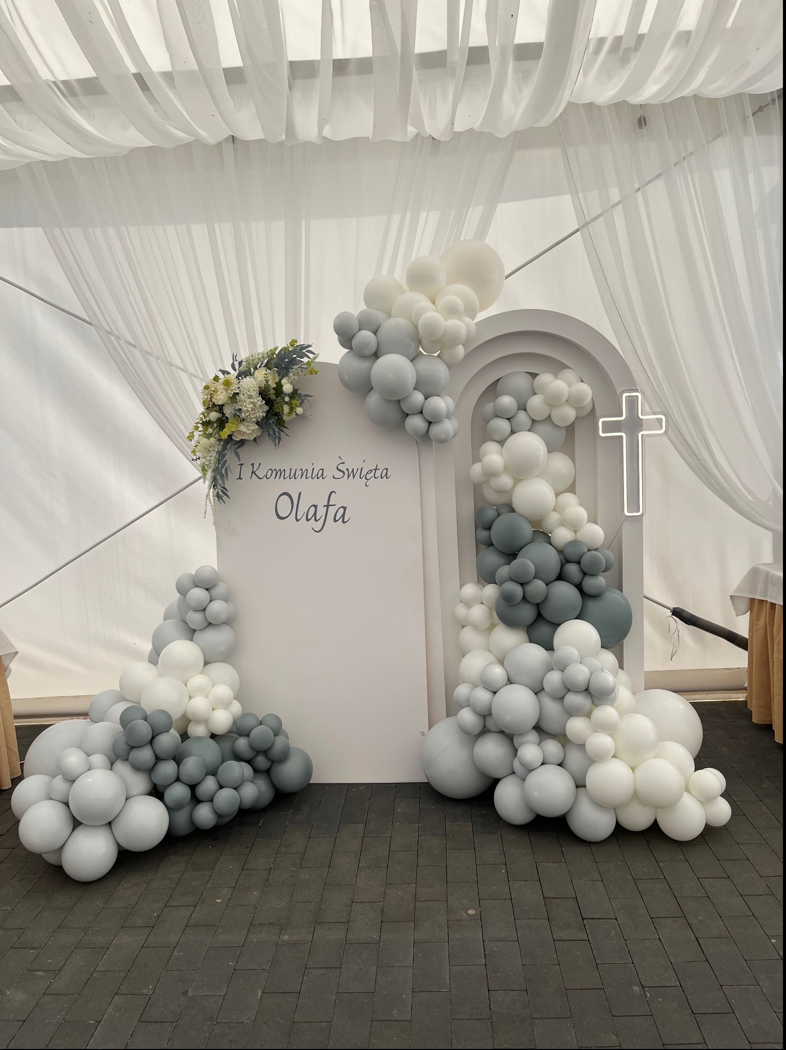Dekoracja na Pierwszą Komunię Świętą Olafa: białe tło z napisem, kwiatowa kompozycja i girlandy z białych i szarych balonów, podświetlony krzyż.