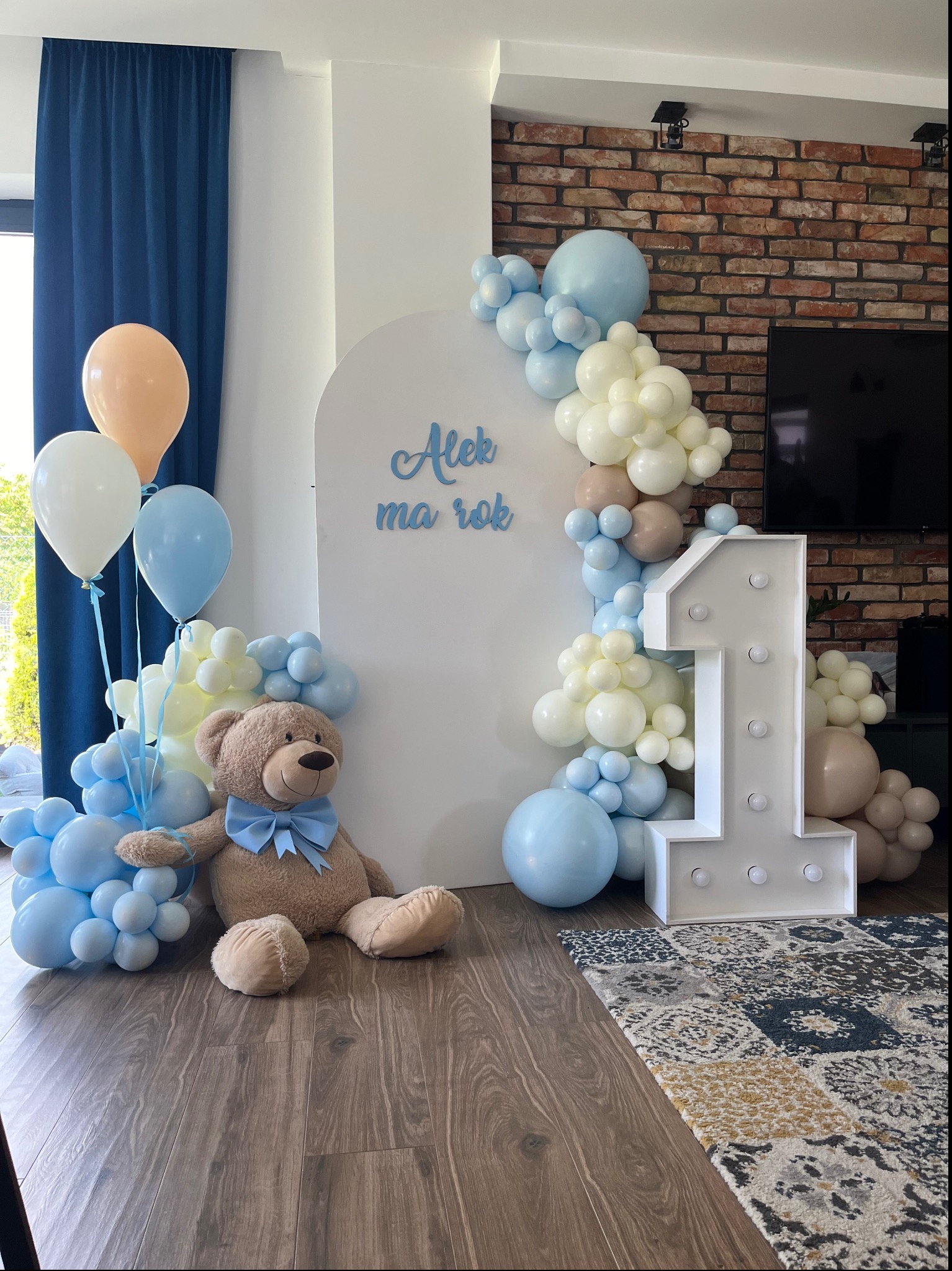 Dekoracja na roczek chłopca: biała ścianka z napisem 'Alek ma rok', girlanda z jasnoniebieskich, białych i beżowych balonów, podświetlana cyfra 1, pluszowy miś z błękitną kokardą i bukiet balonów...