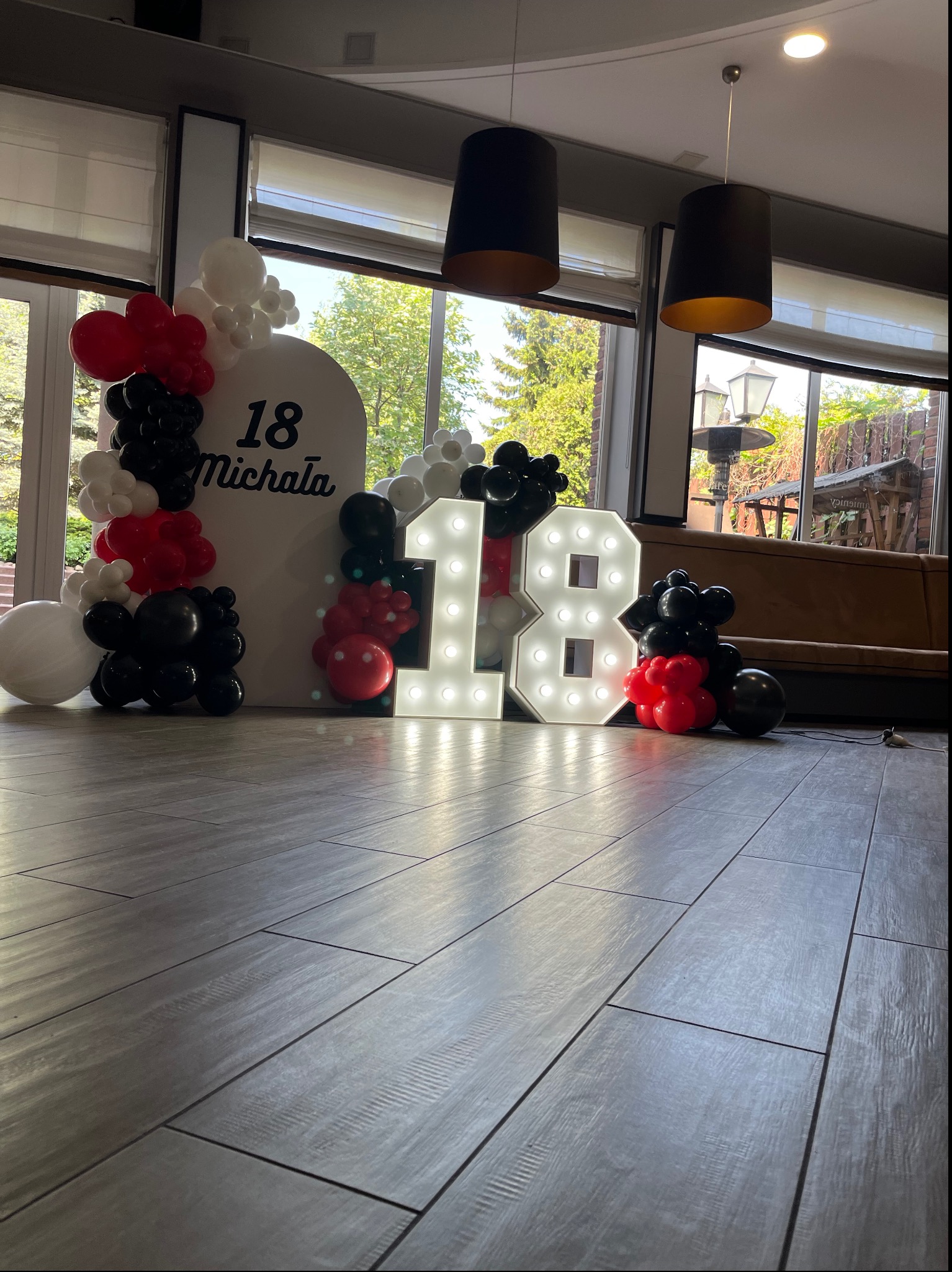 Aranżacja urodzinowa z białym panelem z napisem '18 Michała', podświetlanymi cyframi '18' i girlandami z czarno-biało-czerwonych balonów w pomieszczeniu z dużymi oknami.