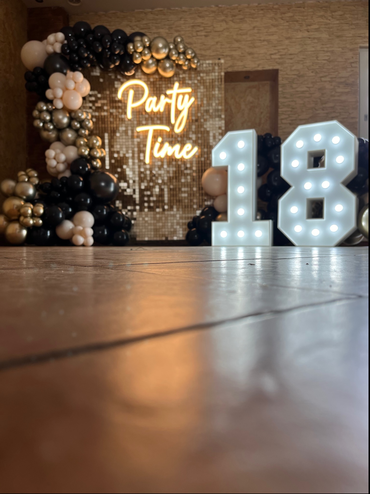 Dekoracja urodzinowa z balonów w odcieniach czerni, złota i beżu, podświetlany napis 'Party Time' na tle błyszczącej ściany i cyfry '18' z żarówkami.