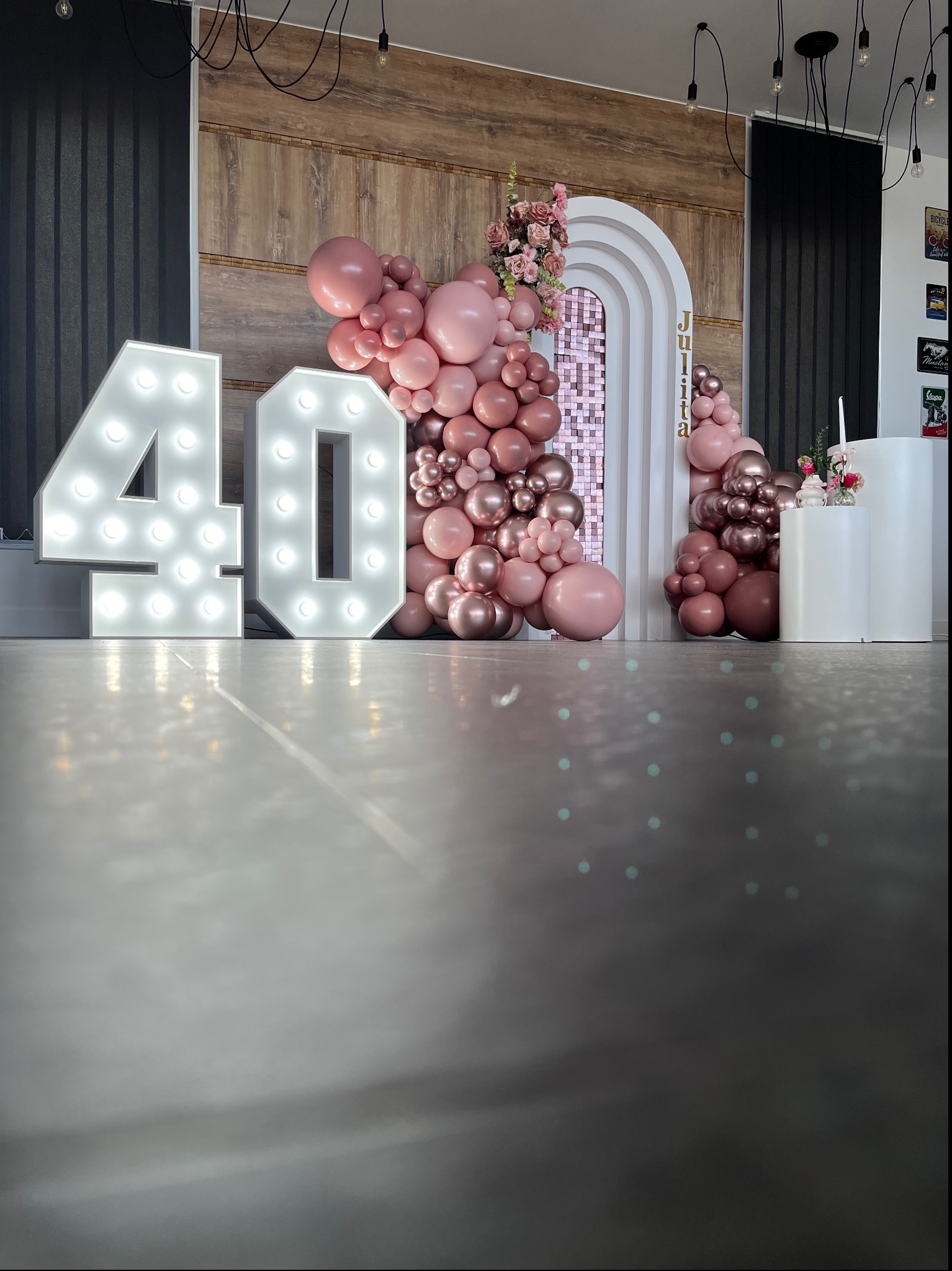 Dekoracja urodzinowa z dużymi podświetlanymi cyframi '40', różowymi balonami, kwiatami i białym łukiem z napisem 'Julita' na tle drewnianej ściany i czarnych zasłon.