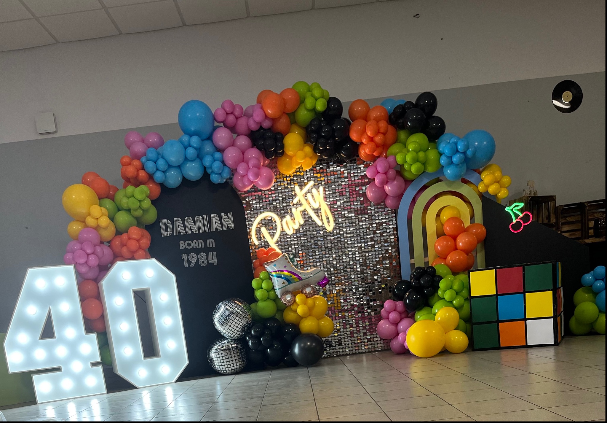 Dekoracja urodzinowa w stylu lat 80. z balonami, neonem 'Party', napisem 'Damian born in 1984', podświetlanymi cyframi '40', rolką i kostką Rubika.