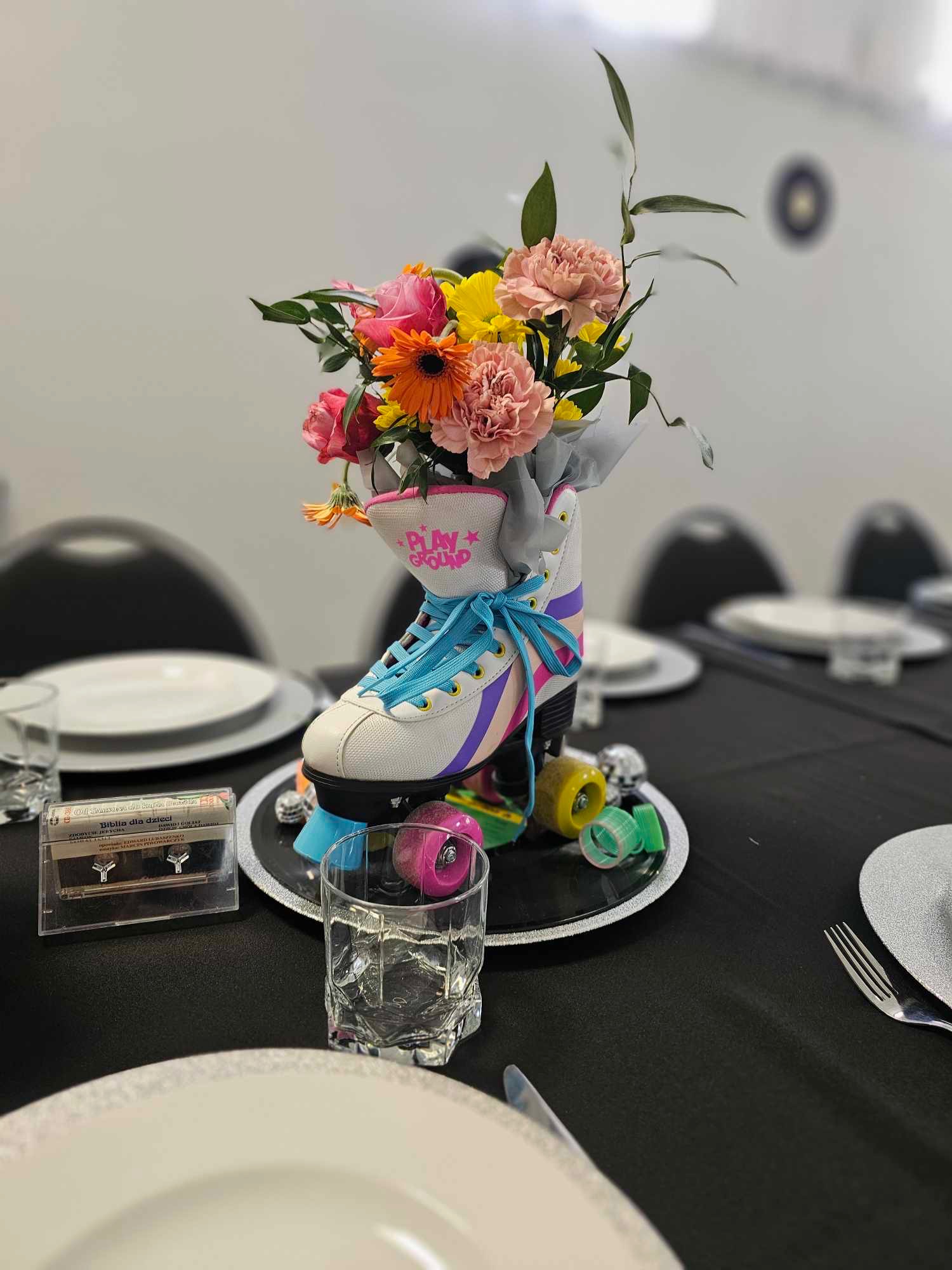 Dekoracja stołu z rolką w stylu retro jako wazonem na kwiaty, otoczona kolorowymi kółkami i kasetą magnetofonową 'Biblia dla dzieci', na czarnym obrusie.