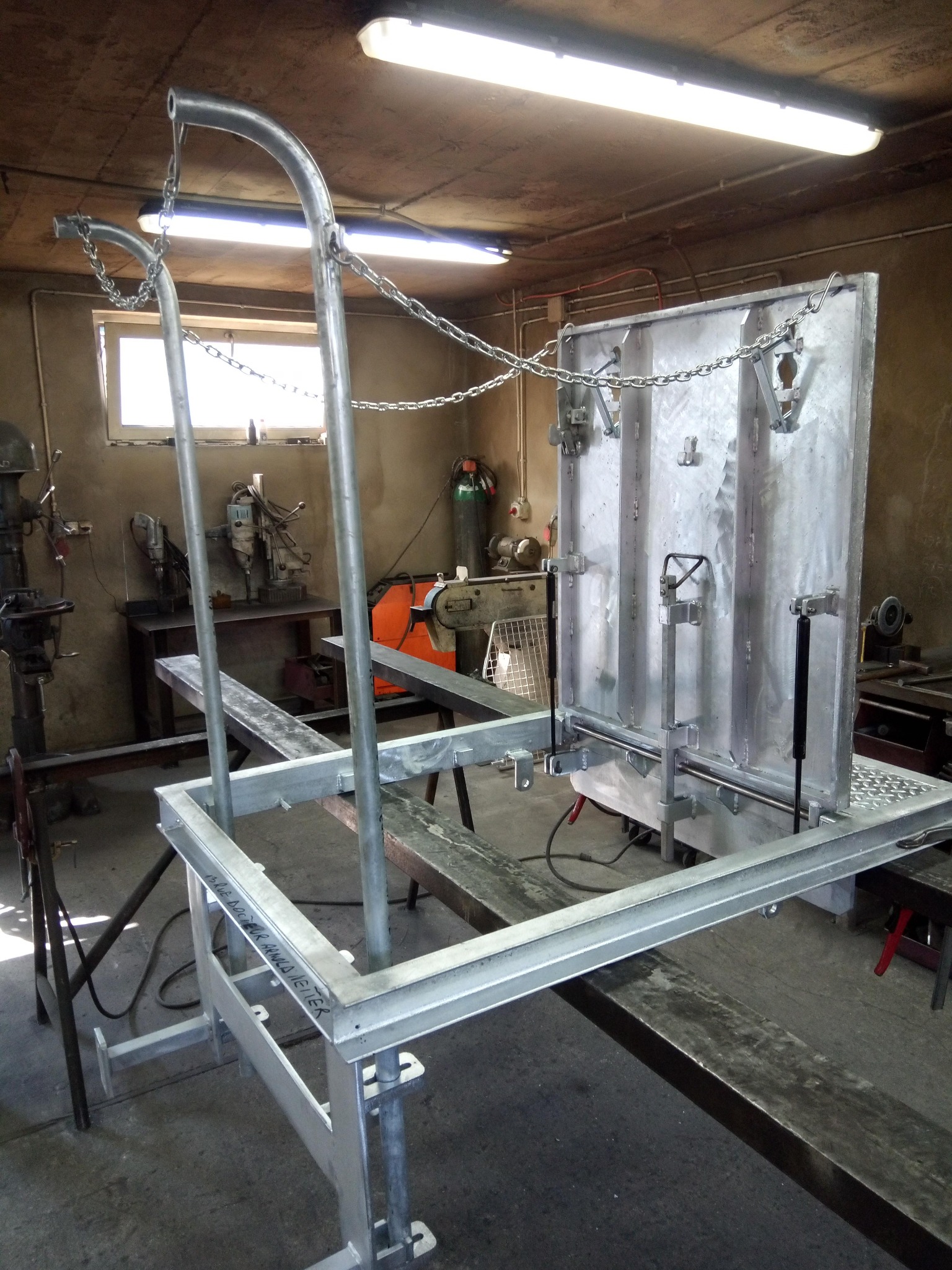 Konstrukcja metalowa z włazem, zawieszona na łańcuchach w warsztacie, widoczne narzędzia i spawarka w tle.