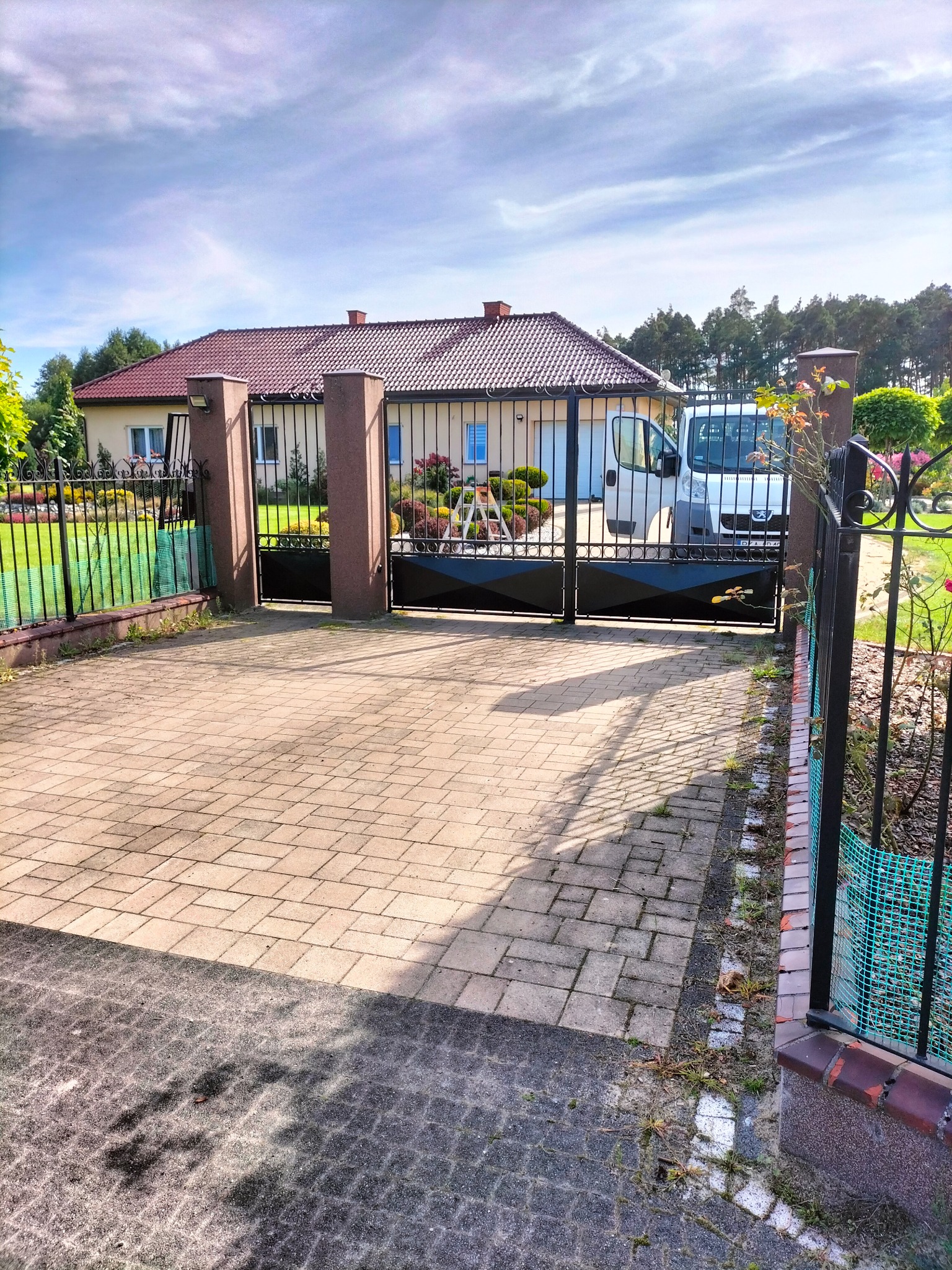 Czarna brama wjazdowa z metalowymi słupkami na posesję z domem jednorodzinnym w tle, na podjeździe z kostki brukowej stoi biały samochód dostawczy.