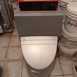 InstalTechSerwis Jakub Pachulski - Kompaktowy piec kondensacyjny Viessmann z czerwonym panelem sterowania, widoczne rury instalacyjne i wiadra z materiałami budowlanymi w tle.