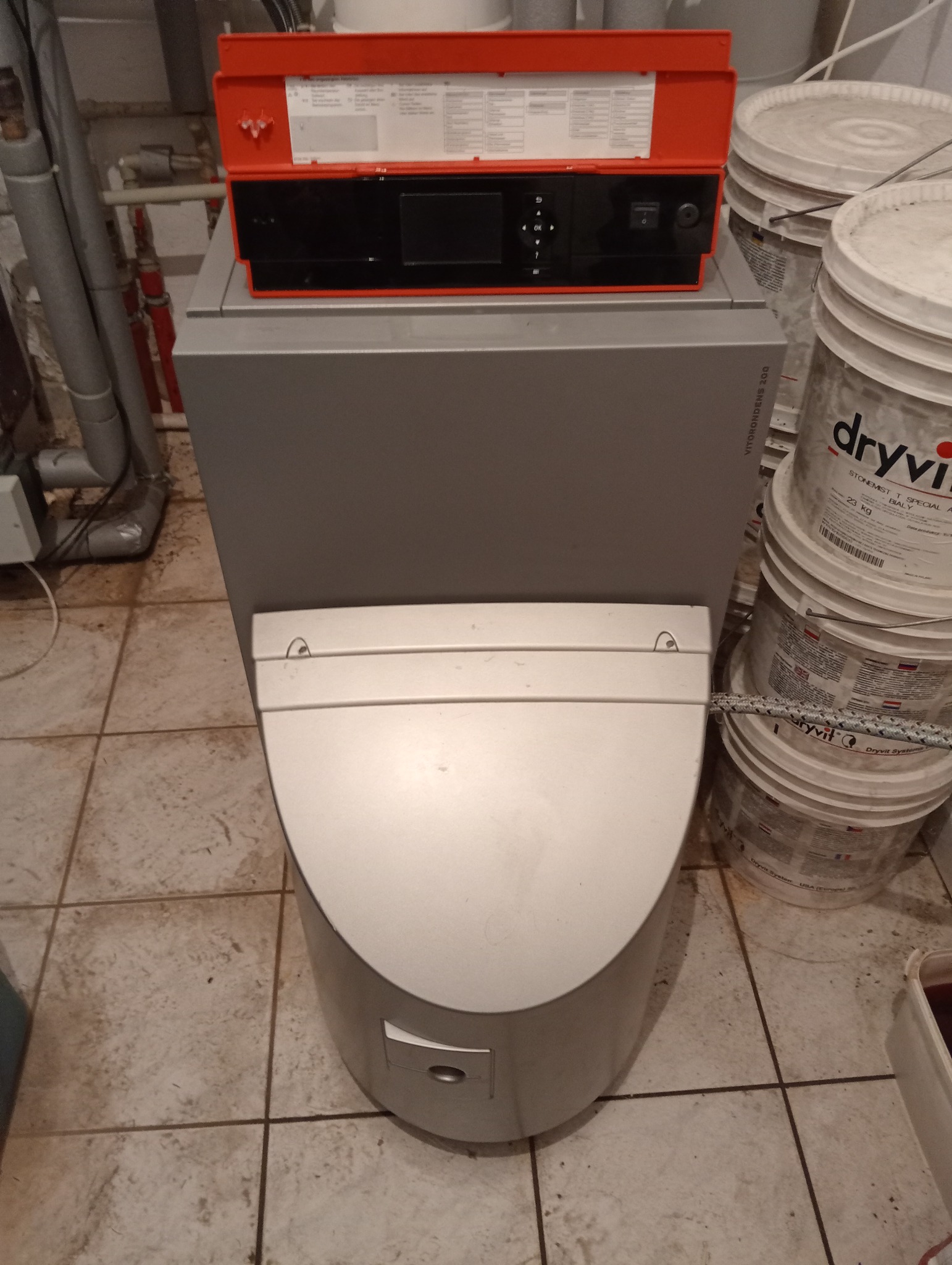 Kompaktowy piec kondensacyjny Viessmann z czerwonym panelem sterowania, widoczne rury instalacyjne i wiadra z materiałami budowlanymi w tle.