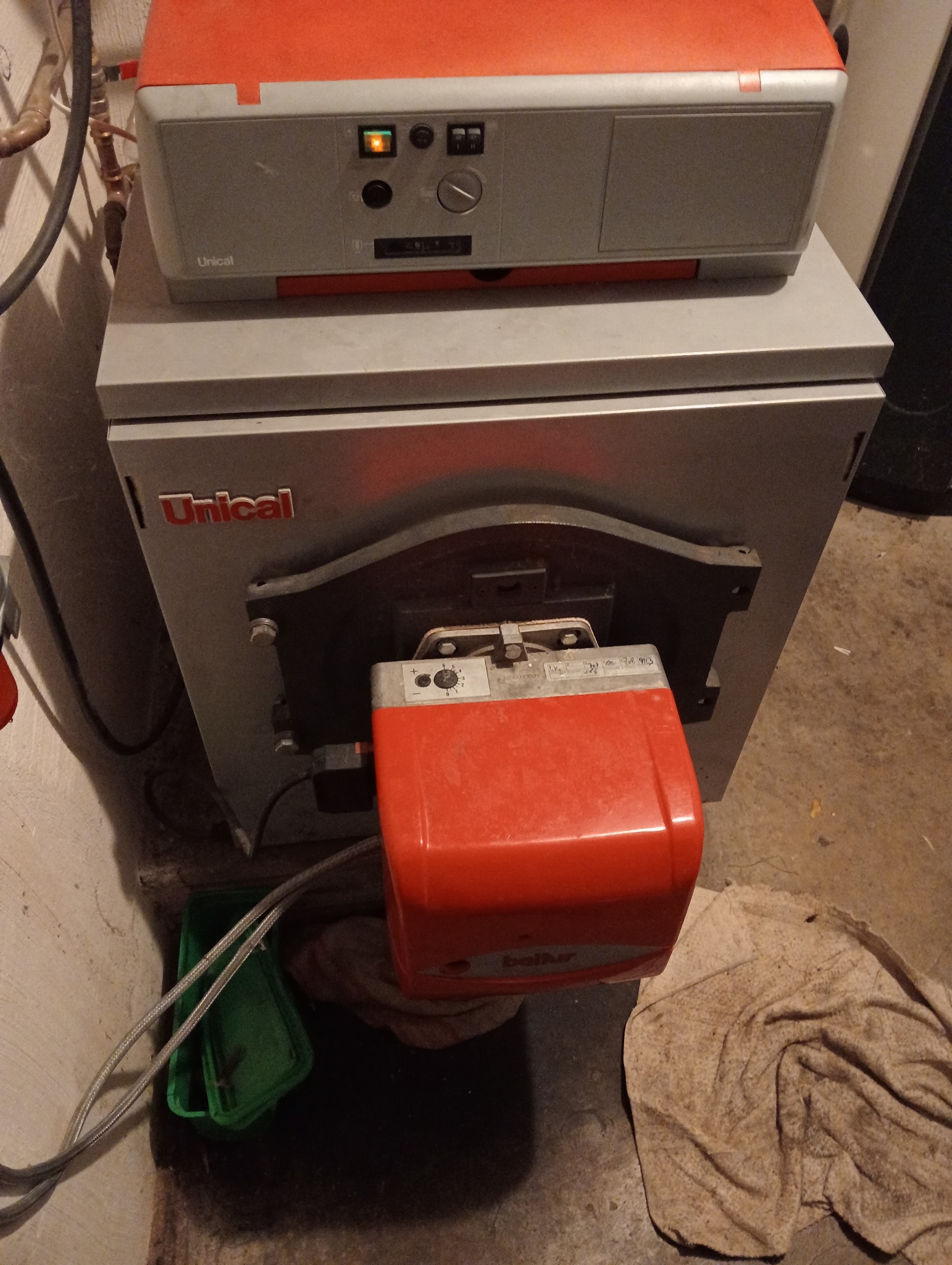 Szary piec CO marki Unical z pomarańczowym palnikiem Baltur, widoczny zielony pojemnik i brudna ściereczka na betonowej podłodze.