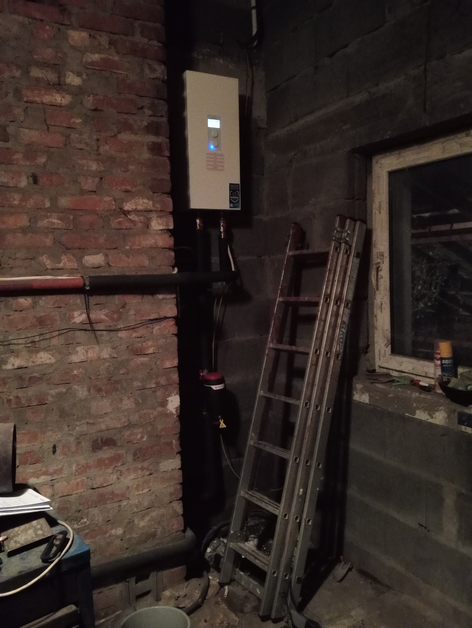 Nowy piec gazowy zamontowany na ścianie z cegły, obok drabina oparta o ścianę betonową, widoczne rury i kable podłączeniowe.