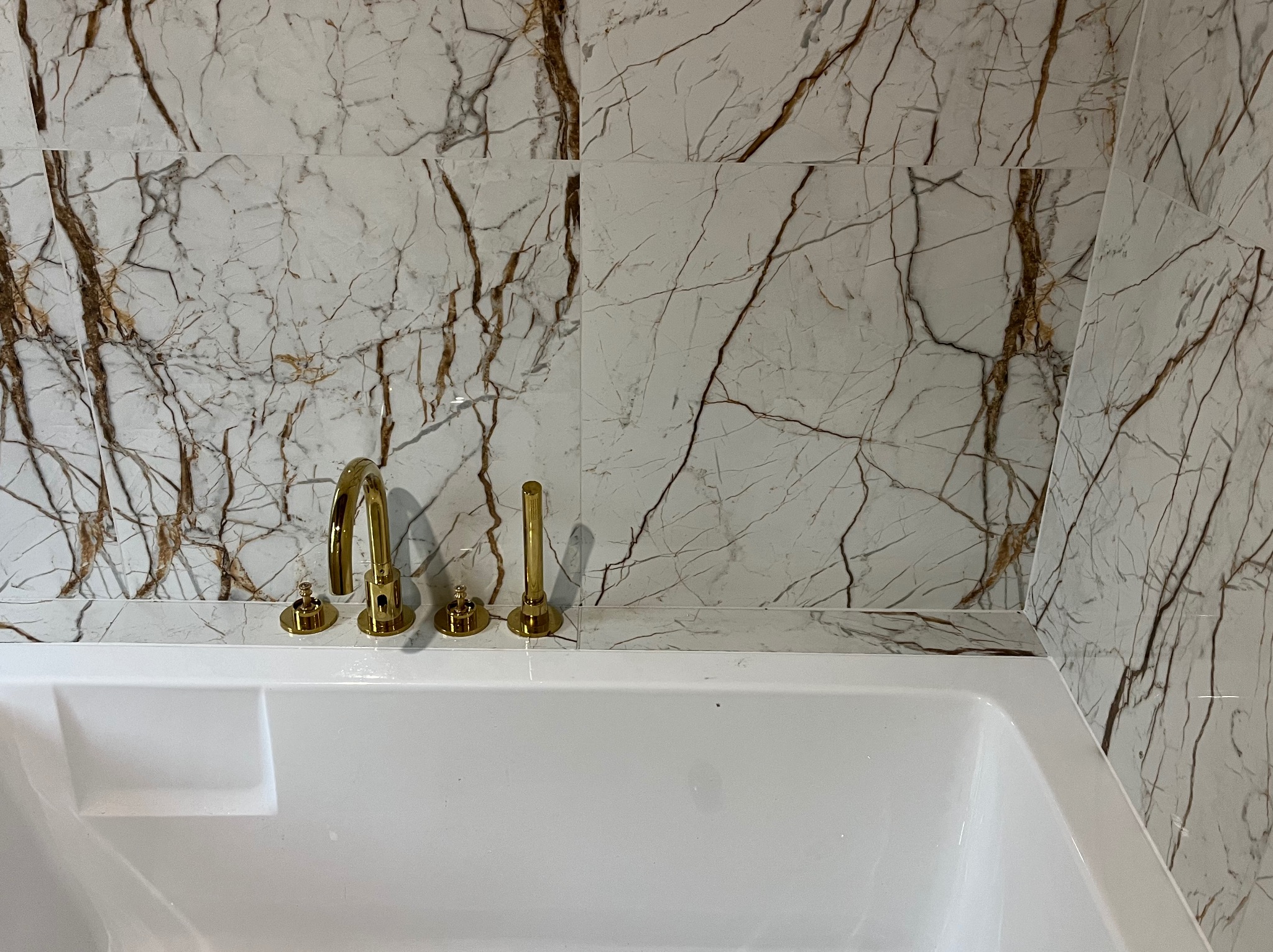 Biała wanna z złotą armaturą na tle ściany wyłożonej płytkami imitującymi marmur z brązowymi żyłami.