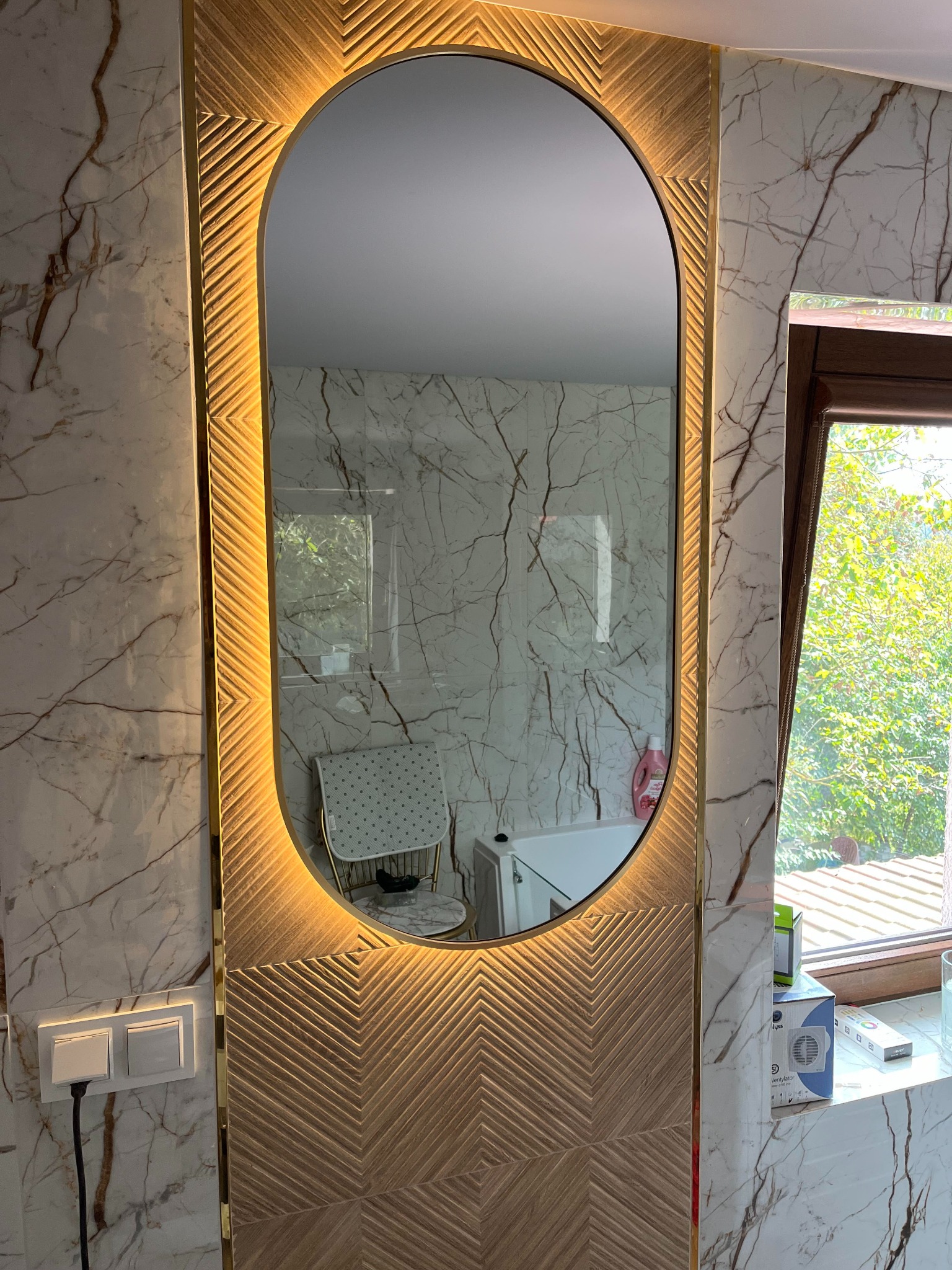 Owalne lustro z podświetleniem LED i dekoracyjną ramą na ścianie wyłożonej marmurowymi płytkami, odbijające fragment łazienki z krzesłem i oknem.