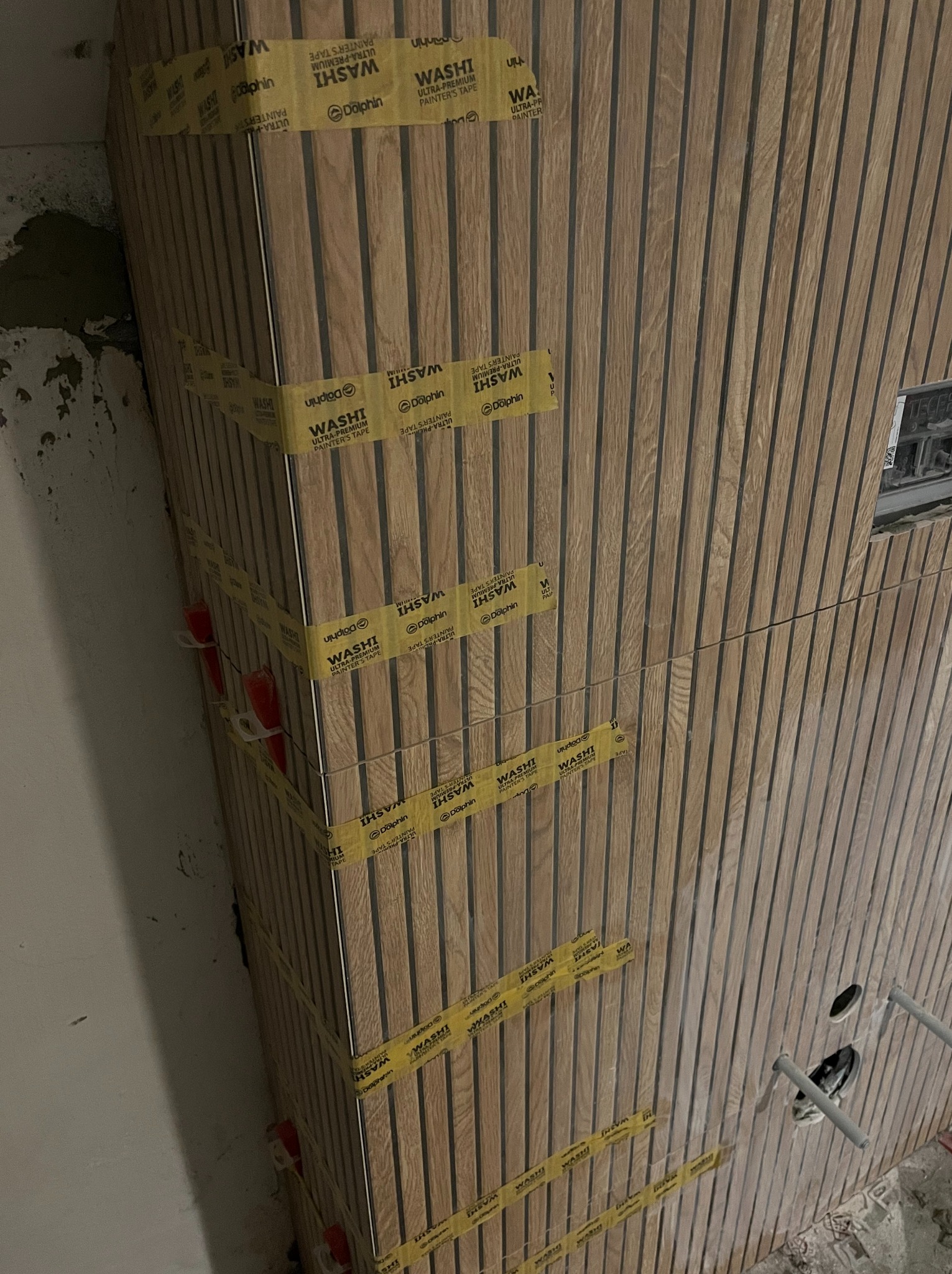 Montaż drewnianych paneli ściennych z użyciem taśmy malarskiej i krzyżyków dystansowych, widoczne otwory na instalacje sanitarne.