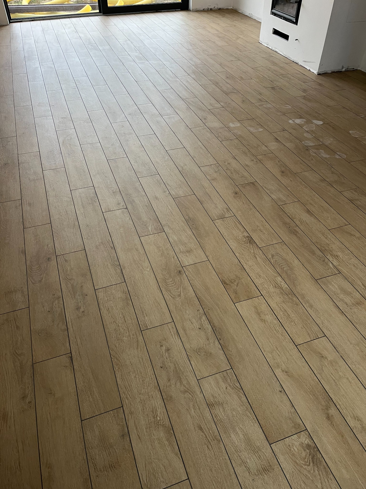 Ułożona podłoga z płytek imitujących drewno w nowym pomieszczeniu, widoczne ślady po budowie i biały kominek z czarnym wkładem.