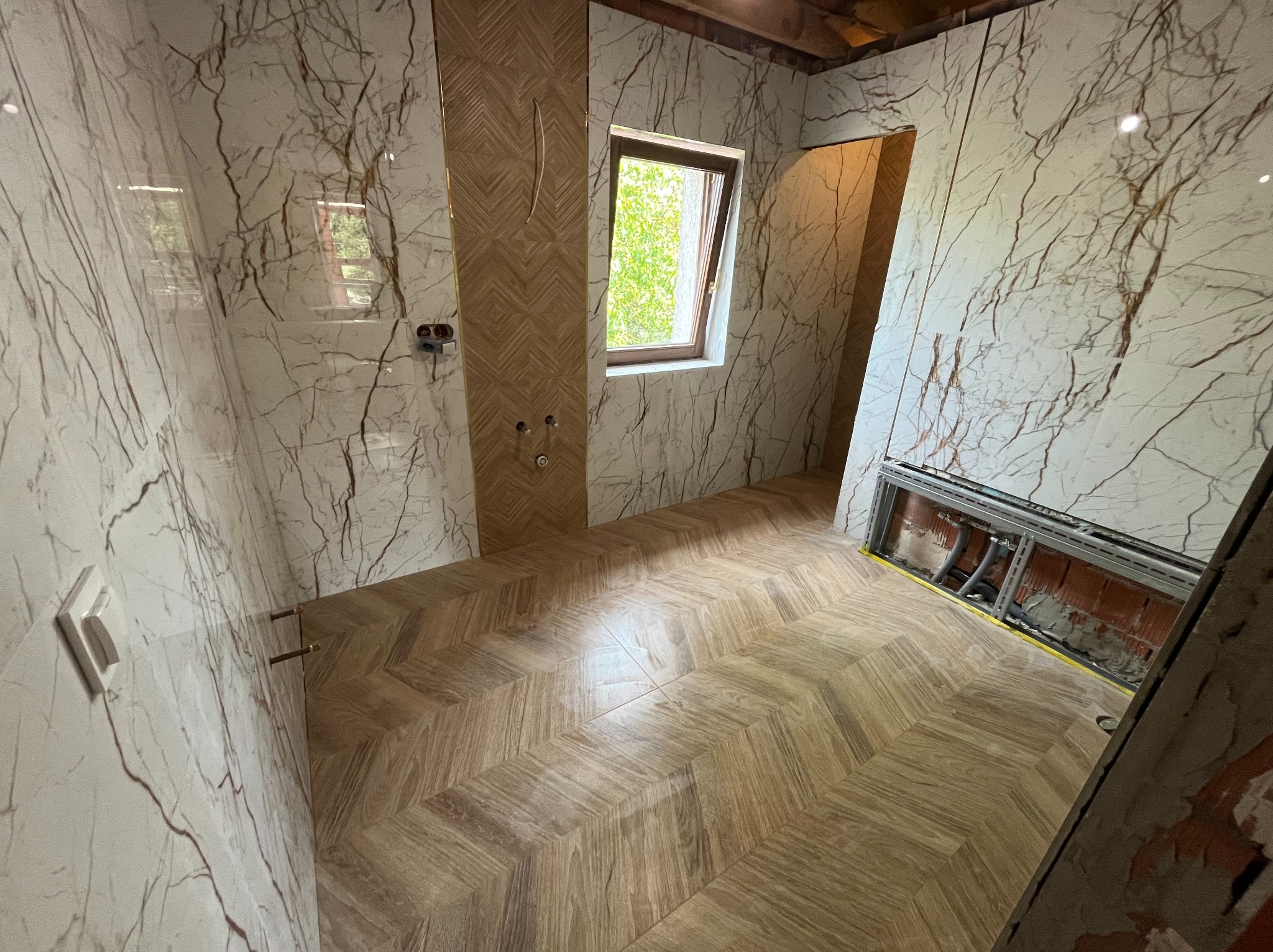 Łazienka w trakcie wykańczania: ściany wyłożone białymi płytami z brązowym żyłkowaniem imitującym marmur, podłoga z drewnopodobnej glazury ułożonej w jodełkę, widoczne instalacje wodne i fragment...