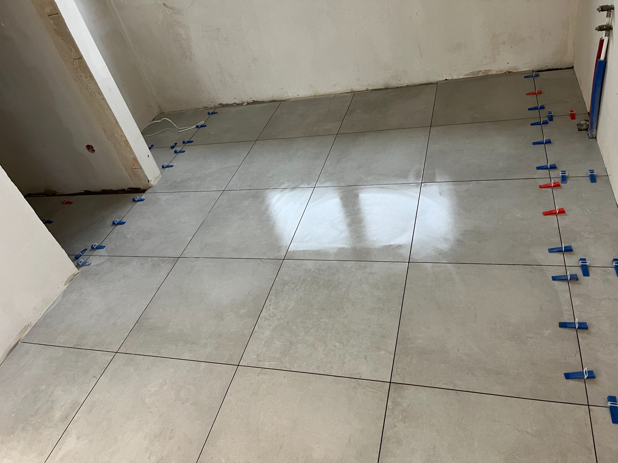 Świeżo ułożone, jasnoszare płytki podłogowe w trakcie poziomowania za pomocą niebiesko-czerwonych klipsów dystansowych na krawędziach, widoczne surowe ściany i instalacje hydrauliczne.