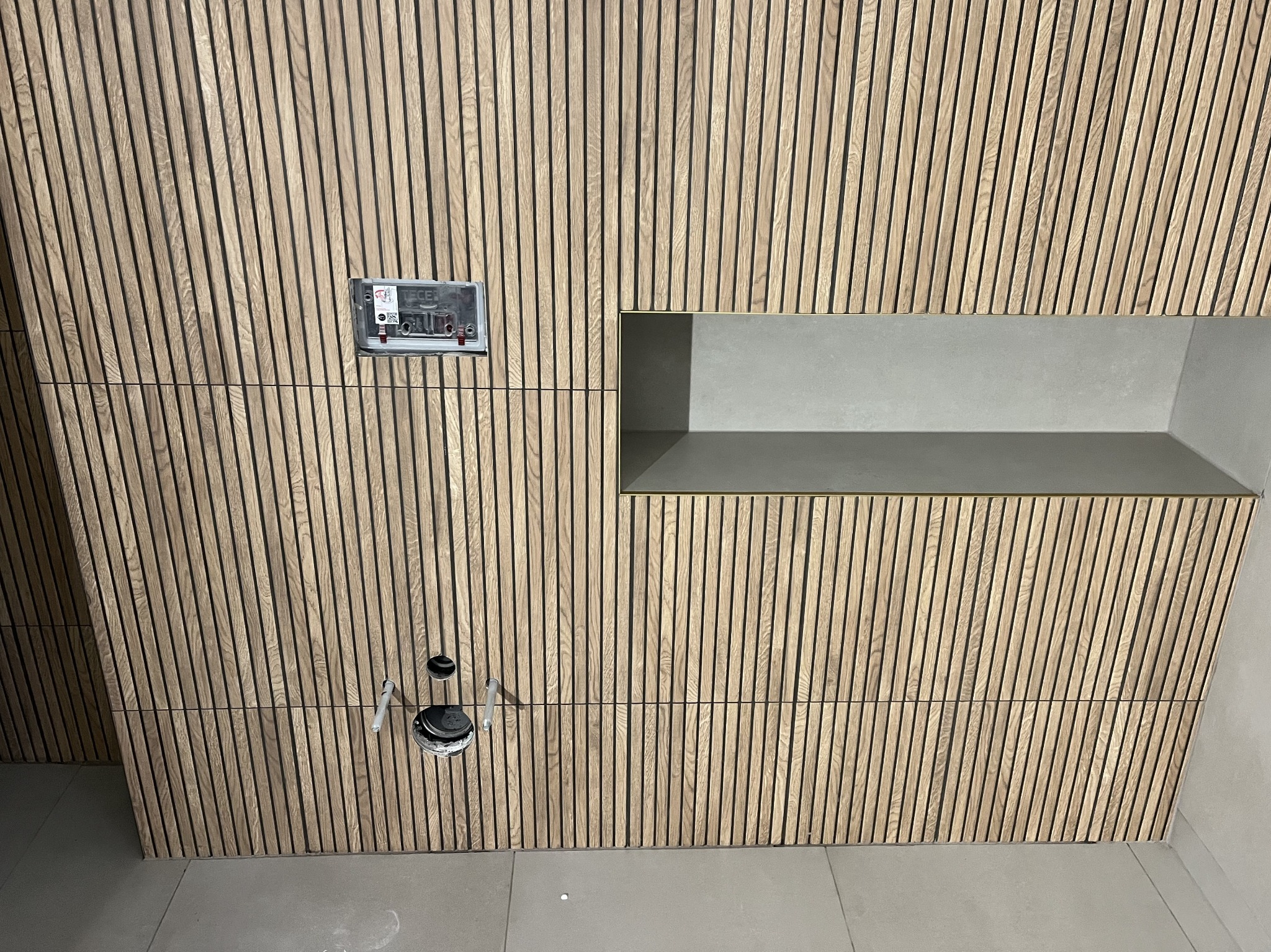Drewniana okładzina ścienna z pionowych listew w łazience, z widoczną wnęką wykończoną płytkami, puszką elektryczną i elementami instalacji hydraulicznej przed montażem armatury.