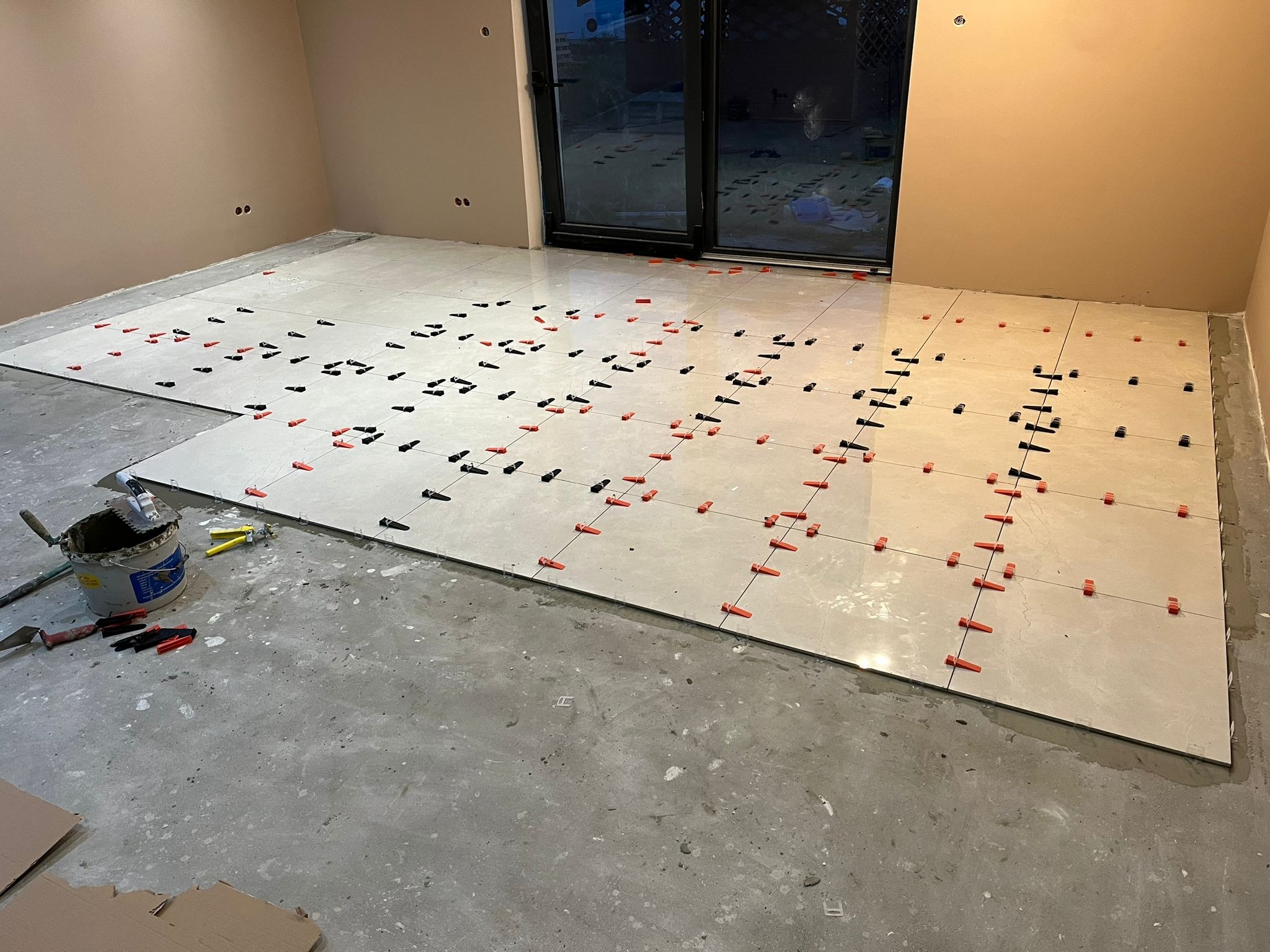 Układanie jasnych płytek podłogowych z wykorzystaniem pomarańczowych i czarnych klipsów poziomujących, widoczne narzędzia glazurnicze i wiadro z zaprawą.