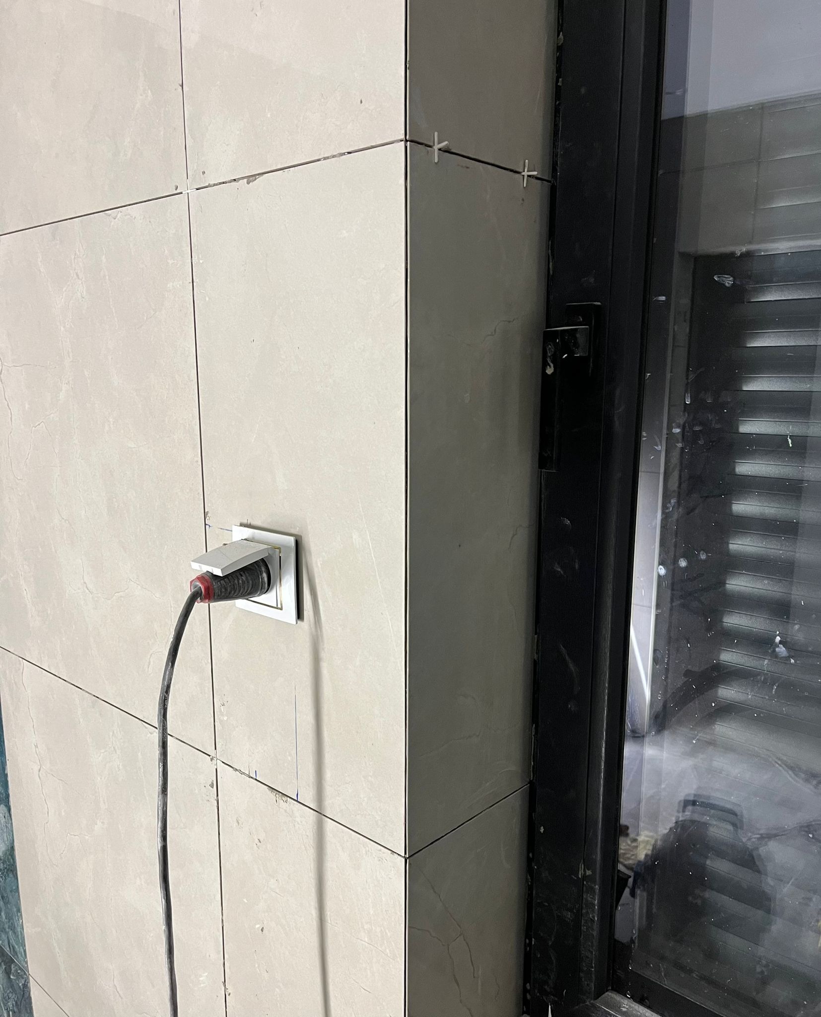 Ściana wyłożona beżowymi płytkami, z widocznymi krzyżykami dystansowymi, gniazdko elektryczne z podłączonym kablem oraz fragment czarnej ramy okiennej.