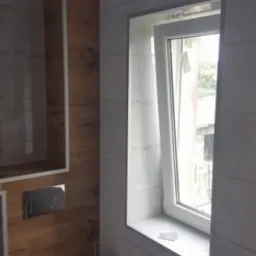 Wykończona łazienka z toaletą, drewnianą zabudową nad spłuczką i oknem z widokiem na zewnątrz, wykończona płytkami w odcieniach szarości.