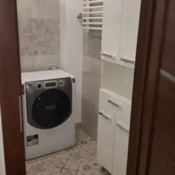 Wyremontowana łazienka z pralką, grzejnikiem drabinkowym i białą szafką, widok zza uchylonych drzwi.