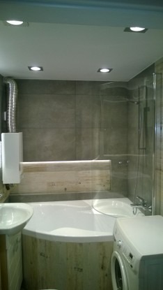 Nowoczesna łazienka z wanną obudowaną drewnem, szarą glazurą, kabiną prysznicową i pralką; oświetlenie sufitowe punktowe.