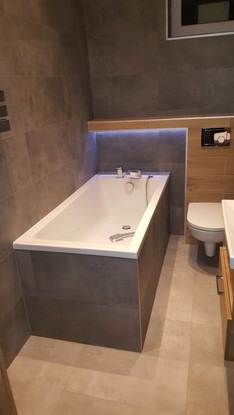 Nowoczesna łazienka z zabudowaną wanną w szarej płytce, podświetlana półka nad wanną, wisząca toaleta z drewnianym panelem.