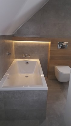 Nowoczesna łazienka na poddaszu z wanną obudowaną szarą płytką, podświetlaną wnęką nad wanną i podwieszaną toaletą.