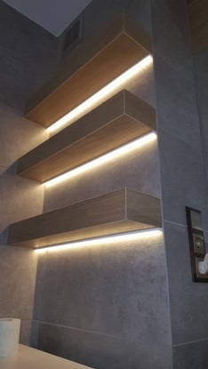 Trzy podświetlane półki z płytek imitujących drewno, zamontowane na szarej ścianie w łazience z widocznym fragmentem toalety.