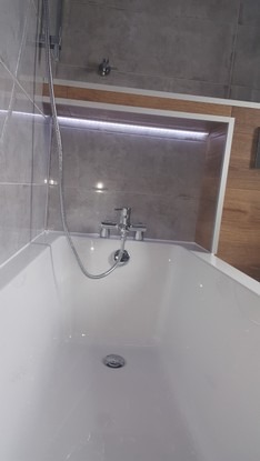 Biała wanna z baterią i słuchawką prysznicową na tle szarej ściany z podświetlaną wnęką, widok z góry.