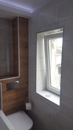 Wykończona łazienka z toaletą, drewnianą zabudową nad spłuczką i oknem z widokiem na zewnątrz, wykończona płytkami w odcieniach szarości.