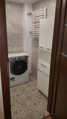 Wyremontowana łazienka z pralką, grzejnikiem drabinkowym i białą szafką, widok zza uchylonych drzwi.