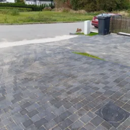 Ułożona kostka brukowa w odcieniach szarości na podjeździe, widoczna studzienka kanalizacyjna, w tle kosz na śmieci, fragment samochodu i domu.