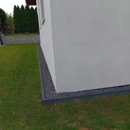 Świeżo otynkowana ściana domu w kolorze złamanej bieli z widocznym oknem, obramowana ciemnoszarym obrzeżem wypełnionym drobnym, szarym kamieniem, przylegająca do trawnika.