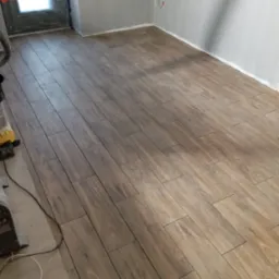 Ułożone płytki podłogowe imitujące drewno w pomieszczeniu z szarymi ścianami, widoczny fragment odkurzacza i zabezpieczone drzwi.